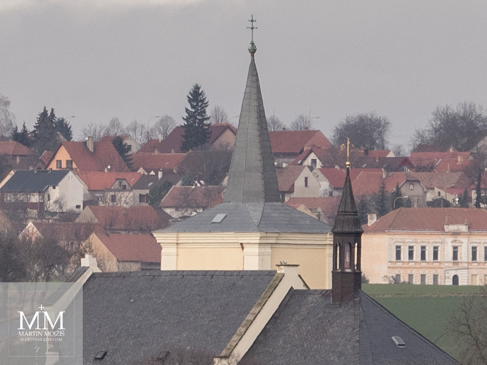 Střecha a věž kostela, v pozadí vesnice. Fotografie vytvořená objektivem Olympus M. Zuiko digital ED 40 - 150 mm 1:2.8 PRO.